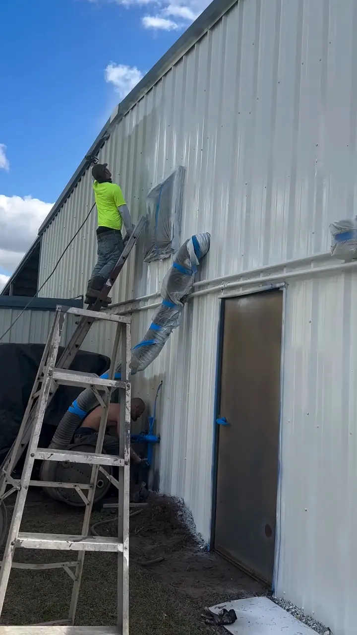 Painter on a ladder working on the wall of a long metal commercial building