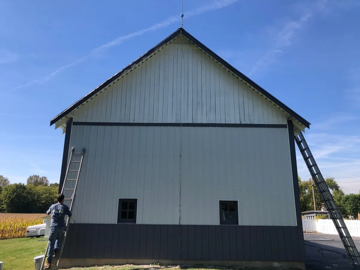 Painter on a ladder at a barn painted light gray with dark gray trim and lower wall