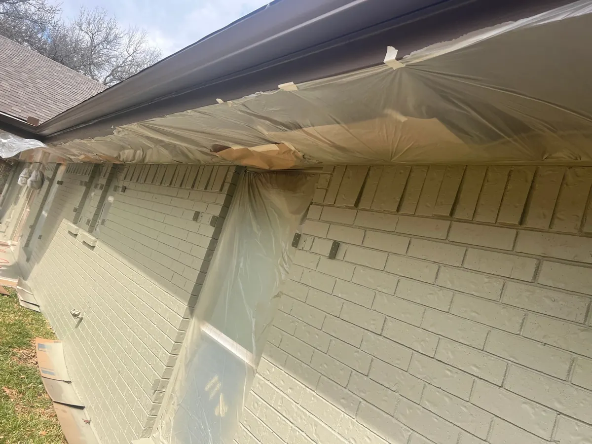 Brick house exterior with the eaves and windows masked in plastic sheeting and tape before painting